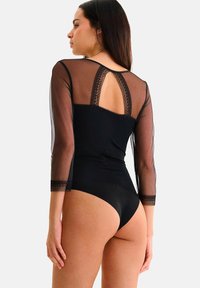 Body noir avec des manches longues transparentes, des accents en dentelle et un design avec ouverture en cœur au dos. Texture lisse avec une silhouette ajustée et une jambe haute.