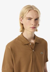 Hnědé polo tričko z texturované látky s dvouknoflíkovou légou a vyšívaným logem na levé straně hrudi. Krátké rukávy.
