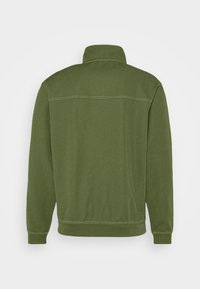 Vihreä fleece-takki korkealla kauluksella, ribbaustetut hihansuut ja kaksi sivutaskua. Sisältää sävysuunnittelua ja sileän pinnan.
