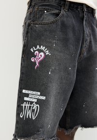 Shorts in denim neri con bordi sfrangiati, macchie di vernice bianca e grafiche stampate che includono "FLAMIN'" e "VERTICAL HORIZON."