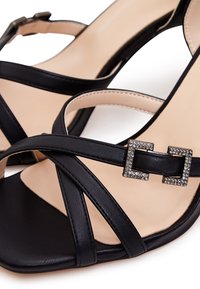 Sandales à talons hauts noires avec des brides croisées, en cuir, dotées d'une boucle carrée ornée de strass et d'une semelle intérieure beige lisse.