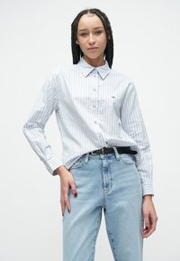 Niebiesko-biała koszula na guziki w paski, wykonana z bawełny, z długimi rękawami, kołnierzykiem i małym logo na lewym piersi. Noszona z jasnymi dżinsami.