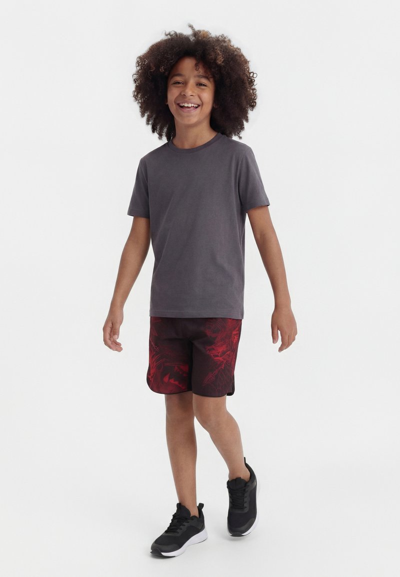 Χαμογελαστό παιδί με σγουρά μαλλιά που περπατά, φοράει σκουρόγκρι μπλουζάκι, κόκκινα και μαύρα σορτς με σχέδια και μαύρα αθλητικά παπούτσια, σε λευκό φόντο.