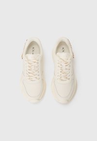 Cream-colored sneakers with a textured upper, lace-up design, gold-tone accents, and a chunky sole. Features branding on the tongue.