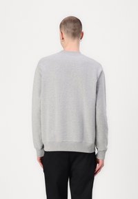 Personne aux cheveux courts et portant des boucles d'oreilles, vêtue d'un pull uni gris clair à manches longues et d'un pantalon noir, vue de dos sur fond blanc.