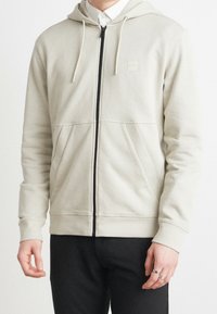 Felpa grigia chiaro con zip in tessuto morbido, dotata di cappuccio con cordoncino, caratterizzata da una zip nera e due tasche frontali. Design semplice e privo di ornamenti.