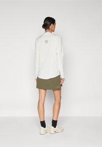 Λευκό πουκάμισο με μακριά μανίκια και μικρό λογότυπο στο πίσω μέρος, συνδυασμένο με ελιά-πράσινη φούστα. Το μοντέλο φοράει μπεζ αθλητικά και σκουρόχρωμες κάλτσες.