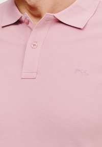 Jasnoróżowa koszulka polo wykonana z teksturowanej tkaniny, z kołnierzykiem, dwiema guzikami i haftowanym logo w dolnym rogu.