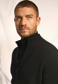 Czarna rozpinana bluza z wysokim kołnierzem, prążkowaną fakturą i dopasowanym krojem. Miękka tkanina na jasnym tle.