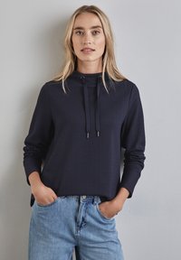 Granatowy, prążkowany hoodie z długim rękawem, z wysokim dekoltem i regulowanymi sznurkami, w połączeniu z jasnoniebieskimi dżinsami.