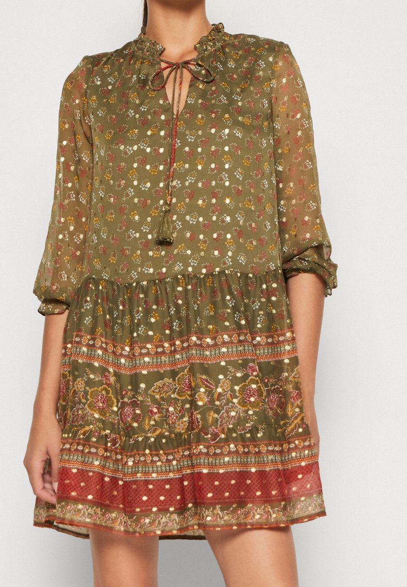 Femme portant une robe fleurie vert olive à manches longues, col froncé, lien à l'avant et ourlet à motifs multicolores au-dessus du genou.
