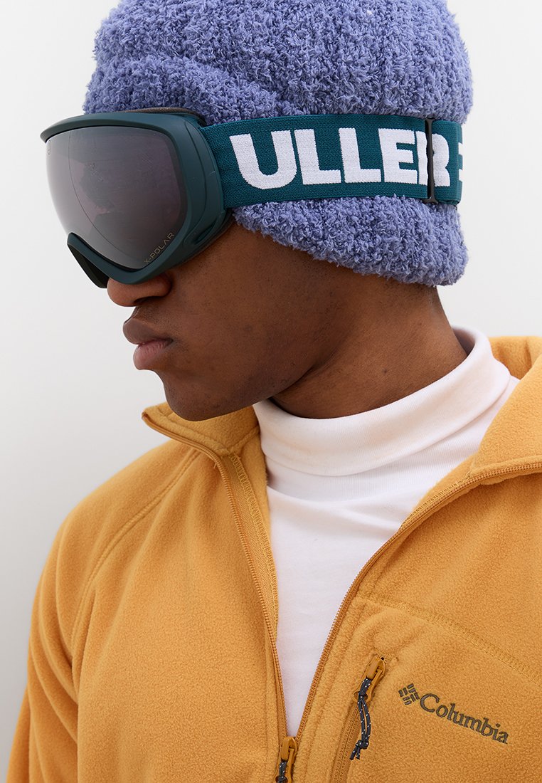 Skibril met een donkere lens en een groene band, gedragen over een paars gestructureerd beanie, gecombineerd met een felgele fleecejas en een witte coltrui.