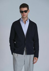 Veste en cardigan marine avec deux poches avant, fabriquée dans un matériau doux. Portée par-dessus une chemise à carreaux bleu clair, assortie à un pantalon de couleur claire.