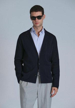 Navy cardigan jacket with two front pockets, made of soft material. Worn over a light blue checkered shirt, paired with light-colored trousers.