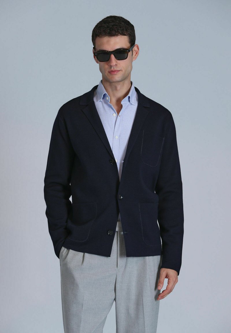 Veste en cardigan marine avec deux poches avant, fabriquée dans un matériau doux. Portée par-dessus une chemise à carreaux bleu clair, assortie à un pantalon de couleur claire.