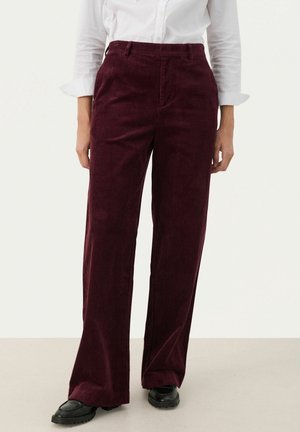 Kobieta nosząca bordowe, szerokie spodnie sztruksowe z wysokim stanem, w połączeniu z czarnymi butami i białą koszulą z długim rękawem i podwiniętymi mankietami.