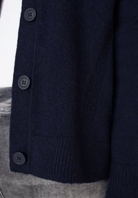 Marineblå cardigan i mjukt stickat material, med sex runda svarta knappar och texturerad ribbing längs kanterna.