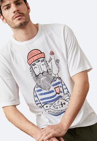Camiseta blanca de algodón con un colorido diseño ilustrado de un hombre barbudo con una camisa a rayas, acompañado de un pájaro y una insignia en el pecho.