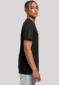 Schwarzes Baumwoll-T-Shirt mit kurzen Ärmeln, Rundhalsausschnitt und lockerer Passform, getragen von einem Modell, das im Profil vor einem neutralen Hintergrund steht.