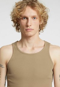 Brun ribbestrikket singlet med rund hals. Stoffet er glatt og tettsittende, med en subtil tekstur. Modellen har krøllete hår og øredobber.