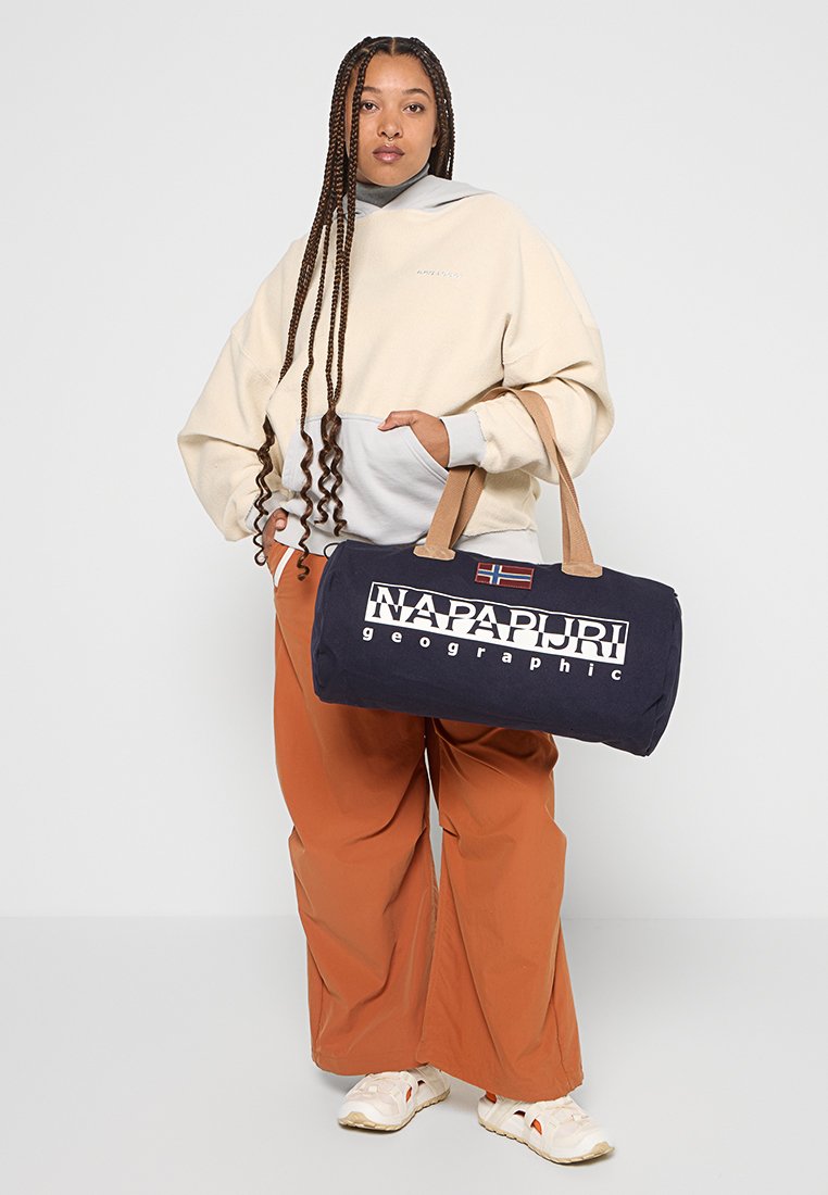 Osoba z długimi warkoczami nosząca beżową bluzę z kapturem i szerokie rudawe spodnie, trzymająca granatową torbę podróżną Napapijri z jasnobrązowymi uchwytami.