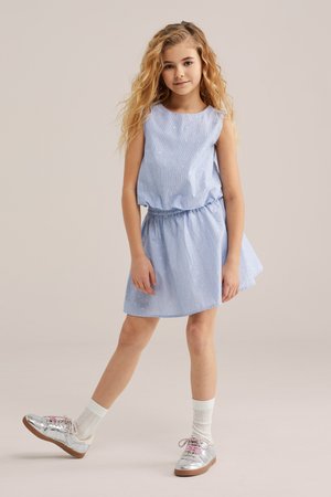 Pikkade lokkis juustega tüdruk, kes kannab helesinist varrukateta kleiti ja hõbedasi tossusid, seistes ühe jalaga välja sirutatud.