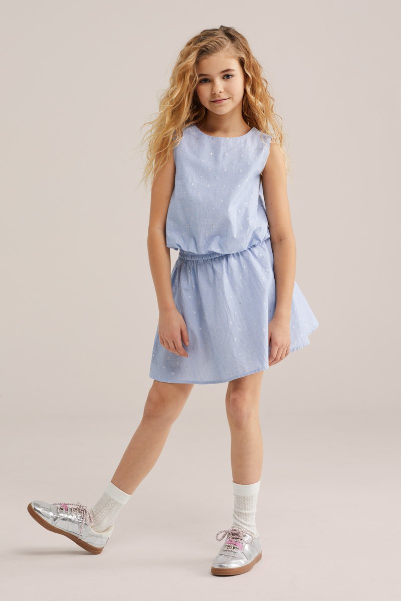 Mädchen mit langen lockigen Haaren, das ein hellblaues ärmelloses Kleid und silberne Turnschuhe trägt und mit einem ausgestreckten Bein steht.