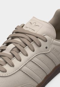 Μπεζ αθλητικό παπούτσι Adidas με μπεζ κορδόνια, τρεις ραμμένες ρίγες στο πλάι και σόλα από καουτσούκ, εμφανίζεται σε κοντινή λεπτομέρεια.