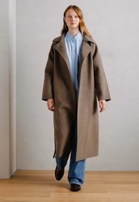 Manteau en laine brun long avec de larges revers, porté sur une chemise à boutons bleu clair et un jean à jambes larges. Texture lisse, design minimaliste.