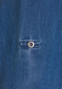 Denimfabric i en mellanblå nyans med en metallknapp med dekorativ kant och kontrasterande sömnad längs fickan.