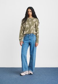 Alyvuogių žalių raštų megztinis su raukšlėta tekstūra, derinamas su šviesiai mėlynomis plačiomis kelnėmis ir šviesiais sportbačiais. Paprastas fonas.