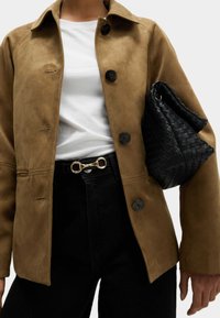 Brun mockajacka med en traditionell krage och svarta knappar, matchad med högmidjade svarta jeans, en vit t-shirt och en vävd svart väska.