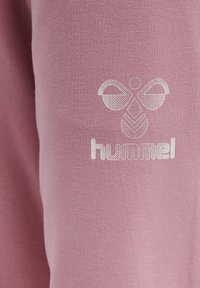 Tissu rose présentant une surface texturée. Un logo blanc avec des formes géométriques et le nom de la marque "hummel" est affiché de manière proéminente.