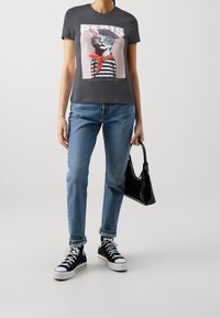 Grå t-shirt med en kattgrafik som bär glasögon och halsduk, tillsammans med ljusblå jeans och svarta högstövlar. Svart handväska ingår.