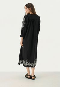 Schwarzes langes Kleid mit weißer Blumenstickerei am Saum und den Ärmeln. Es hat puffige Ärmel und einen lockeren Schnitt, hergestellt aus weichem Stoff.
