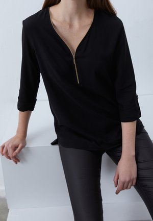 Donna che indossa una maglietta nera con zip e maniche arrotolate, insieme a pantaloni di pelle nera, seduta con una mano appoggiata su una superficie bianca.