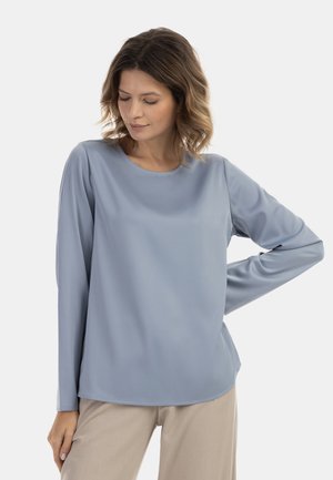 Blusa azzurro chiaro a maniche lunghe in tessuto morbido con scollatura rotonda, vestibilità rilassata e orlo leggermente curvo. Abbinata a pantaloni neutri.