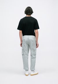 Person med axellångt hår som bär en svart t-shirt, ljusgrå cargobyxor, vita sneakers, står vänd bort från kameran mot en enfärgad vit bakgrund.
