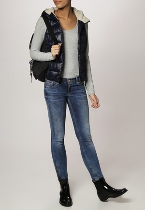 Kobieta ubrana w dopasowane niebieskie dżinsy, czarne błyszczące botki za kostkę, szary top z długim rękawem, granatową kamizelkę puchową z polarową podszewką, niosąca czarny plecak.