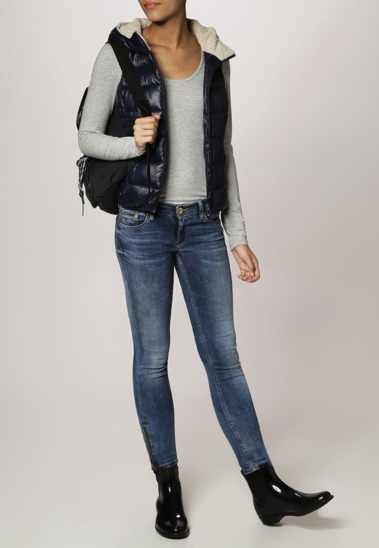 Femme portant un jean bleu ajusté, des bottines noires brillantes, un top gris à manches longues, une veste matelassée bleu marine doublée en polaire, portant un sac à dos noir.