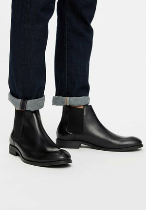 Botines Chelsea de cuero negro con paneles elásticos en los laterales y un acabado suave. Punta redondeada y tacón bajo apilado, combinados con jeans de mezclilla oscura.