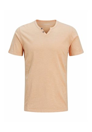 Kortärmad T-shirt i ljus persika, texturerat tyg med V-hals och två knappdetaljer vid kragen. Avslappnad passform, minimal design.