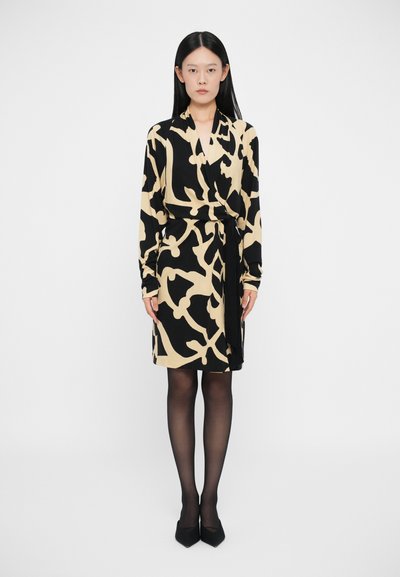 Zwarte en beige wikkeljurk met abstract patroon, lange mouwen en een strik taille, gecombineerd met zwarte hoge hakken en sheer panty.