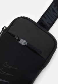 Svart nylonpåse med dragkedja, med en texturerad yta, rundade kanter och ett band med en logotyp. Detaljer i metall är synliga.