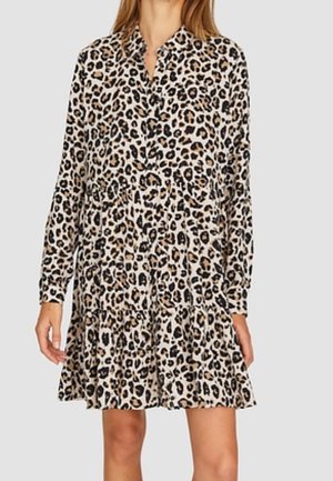 Femme portant une mini-robe à manches longues boutonnée avec un motif léopard beige, noir et marron sur fond uni.