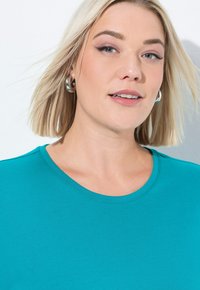 T-shirt di cotone verde acqua con colletto rotondo, caratterizzato da una texture liscia e maniche corte, indossato da una persona con capelli biondi di lunghezza media.