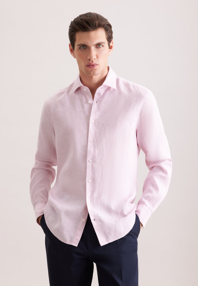 Jeune homme aux cheveux foncés portant une chemise rose pâle à manches longues boutonnée et un pantalon sombre, debout les mains dans les poches.