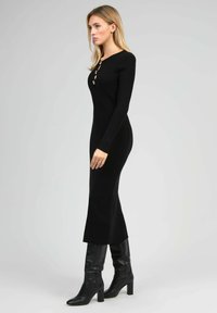 Robe tricot ribbed noire à manches longues et silhouette ajustée. Présente trois accents de boutons au niveau du col ; associée à des bottes noires à hauteur de genou.