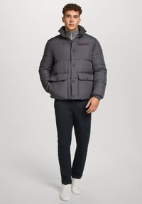 Giacca puffa grigio scuro, con colletto alto, chiusura frontale con bottoni, due grandi tasche e polsini a coste, indossata sopra un dolcevita grigio chiaro.