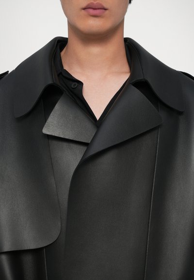 Sort læder trench coat med bred krave, struktureret revers og glat tekstur. Laget over en sort skjorte med knapdetalje.